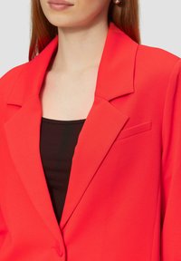 Blazer rouge avec un design ajusté, présentant des revers crantés et une poche poitrine unique. Le tissu semble lisse et légèrement structuré.