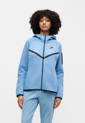 Kobieta z kręconymi włosami ubrana w jasno-niebieską bluzę Nike z suwakiem i pasujące spodnie dresowe, stojąca na jednolitym tle.