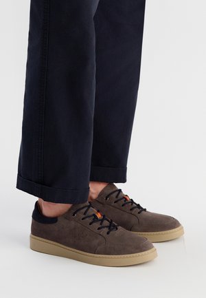 Braune Wildledersneaker mit einer beigen Gummisohle, ausgestattet mit dunkelblauen Schnürsenkeln und einem bunten Akzent an der Zunge. Getragen mit dunklen Hosen.