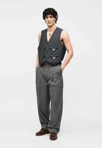 Homme aux cheveux bouclés portant un gilet noir avec quatre poches sur le devant, un pantalon large rayé, des chaussures marron et un collier pendentif, debout les mains dans les poches.