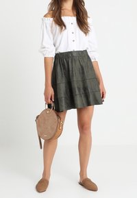Top bianco scollato con bottoni, gonna a strati verde scuro, borsa rotonda color cammello e scarpe slip-on intrecciate marroni.