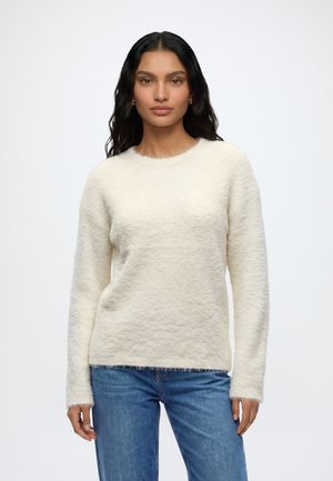 Femme aux longs cheveux foncés portant un pull blanc duveteux à manches longues et un jean bleu, debout devant un fond clair uni.