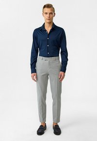 Marineblauwe overhemd met lange mouwen en knopen, grijze op maat gemaakte broek, en donkere instappers. Het overhemd heeft een zachte textuur; de broek is recht gesneden en van lichtgewicht stof.