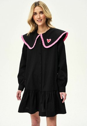 Kobieta ubrana w czarną sukienkę zapinaną na guziki, z dużym kołnierzem obszytym różowym kolorem oraz małym różowym serduszkiem po lewej stronie kołnierza.