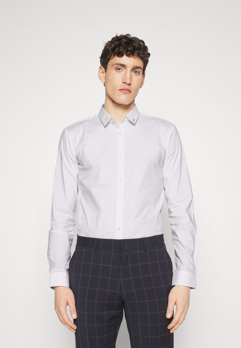 Light gray long-sleeve shirt with a pointed collar, buttoned front, and structured fit. Paired with navy plaid trousers.