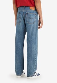 Personne portant un jean bleu droit, une chemise rouge et des baskets bleues et blanches, debout les mains détendues le long du corps.
