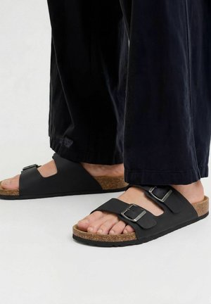 Noge u crnim sandalama s plosnom od pluta, s dva podesiva kopčasta remena i crnim hlačama stoje na bijeloj površini.