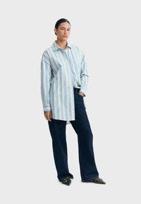 Femme portant une longue chemise à rayures verticales bleues et blanches, un pantalon large sombre et des chaussures noires pointues, debout avec une main dans la poche.