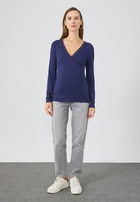 Sötétkék hosszú ujjú felső v-nyakkivágással és csipkés szegéllyel, világosszürke egyenes szárú farmernadrággal és fehér tornacipővel, semleges háttéren.