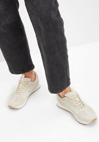 Scarpe da tennis beige in materiale mesh e suede, punta rotonda, design con lacci e suola di gomma, abbinate a pantaloni di jeans neri.