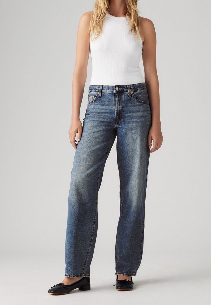 Femme portant un débardeur blanc, un jean bleu taille haute à jambes droites et des ballerines noires, debout devant un fond gris.
