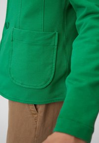 Zelená bavlněná bunda s náprsní kapsou, zapínáním na knoflíky a vyšívanými detaily. V kombinaci se světle hnědými kalhotami. Hladká textura, ležérní design.