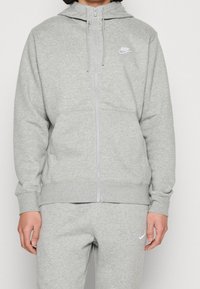 Person som bär en ljusgrå Nike-hoodie och matchande svettbyxor med dragkedja framtill och små vita Nike-loggor på bröstet och på låret.