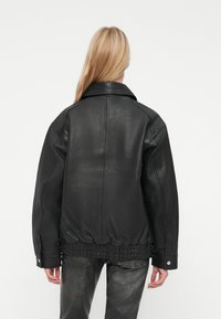 Persoon met lang blond haar, gekleed in een losse zwarte leren blazer en donkergrijze broek, die met de rug naar de kijker staat en de handen ontspannen langs de zijden heeft.