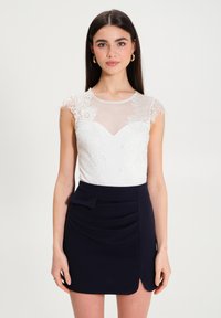 Top bianco in pizzo a maniche corte con corpetto trasparente, abbinato a una gonna blu scuro aderente con plissettatura laterale e dettaglio a fiocco sulla vita.