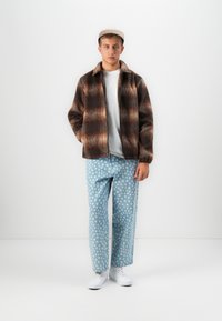 Jonge man in een bruin geruite jasje, witte t-shirt, blauwe broek met stippen, witte sneakers en een beige pet, staand tegen een witte achtergrond.