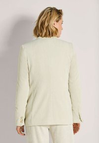 Vrouw met kort blond haar, gekleed in een crèmekleurige corduroy blazer en bijpassende pantalons, met haar rug naar de camera, tegen een effen lichte achtergrond.