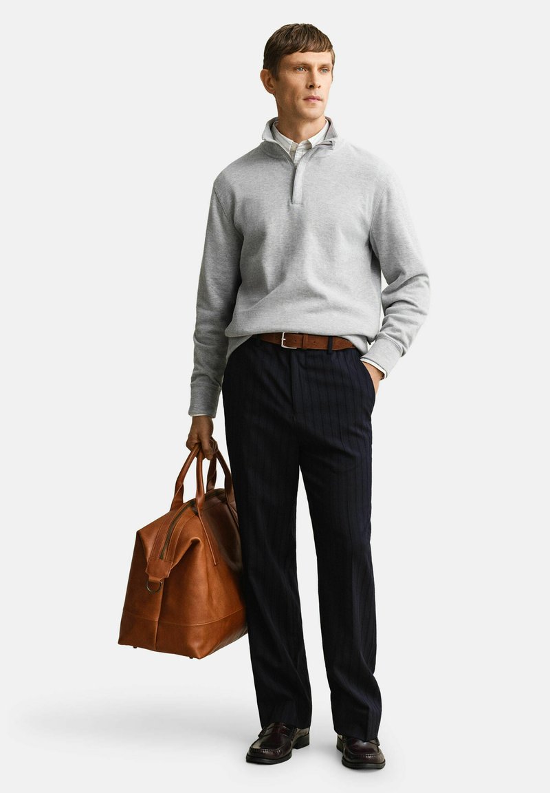 Mann trägt einen hellgrauen Pullover, ein weißes Hemd, dunkle Nadelstreifenhose und braunen Gürtel, hält eine große braune Leder-Reisetasche.