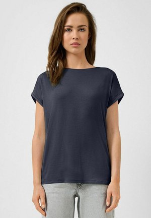 Jonge vrouw met bruin haar, gekleed in een donkerblauw T-shirt met korte mouwen en lichtgrijze spijkerbroek, staand tegen een effen lichte achtergrond.