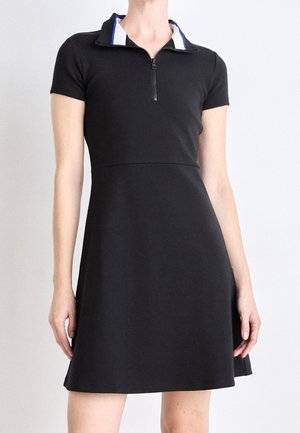 Femme portant une robe noire à manches courtes avec une fermeture éclair devant et un col blanc et bleu, debout devant un fond blanc uni.