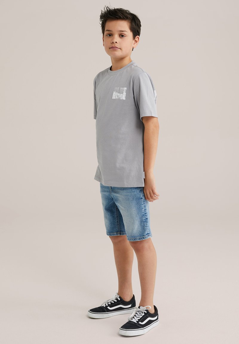 Pelēka kokvilnas T-krekls ar īsām piedurknēm un „Blue Ridge” grafiku, apvienots ar gaiši ziliem džinsu šortiem un melniem sporta apaviem.