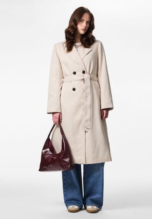 Mujer con un abrigo largo beige con cinturón, vaqueros anchos azules, zapatillas beige, sosteniendo un bolso de cuero burdeos oscuro sobre un fondo blanco.