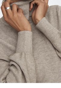 Maglione beige a collo alto realizzato in morbido knit testurizzato. Presenta maniche lunghe con polsini a costine, che mettono in mostra mani adornate da un anello d'argento.