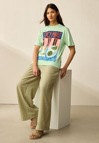Ljusgrön t-shirt med ett färgglatt motiv som visar drycker och lime, kombinerad med lösa, rutiga beige byxor och sandaler.