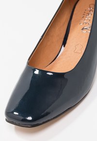 Chaussure plate pour femme en cuir verni bleu foncé avec bout carré et semelle intérieure marron clair, montrant une partie du nom de la marque et un tampon indiquant le matériau en cuir.