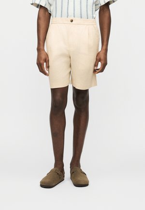 Person wearing beige knee-length shorts, striped short-sleeve shirt, and brown slip-on shoes standing against a plain background.