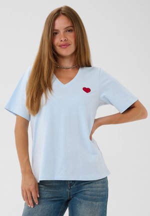 Jeune femme aux longs cheveux bruns porte un t-shirt bleu clair à col en V avec un petit cœur rouge sur la poitrine et un jean bleu, debout, main sur la hanche.