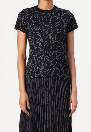 Kobieta w czarnym topie z krótkim rękawem z dużymi konturami kwiatów i plisowaną ciemną spódnicą z abstrakcyjnymi wzorami.
