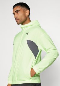 Veste à capuche vert clair avec logo Puma noir, dotée de panneaux latéraux en maille grise et de poignets élastiques. Texture lisse, design ajusté pour le sport.