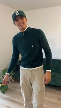 Tmavozelený sveter na zips s bielym golierom, v kombinácii so svetlobéžovými nohavicami typu cargo. Na ľavej strane hrude malý nápadný logo.