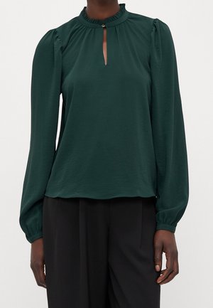 Blouse à manches longues vert foncé avec une petite ouverture en forme de goutte sur le devant et un col à volants, portée avec un pantalon noir ample.