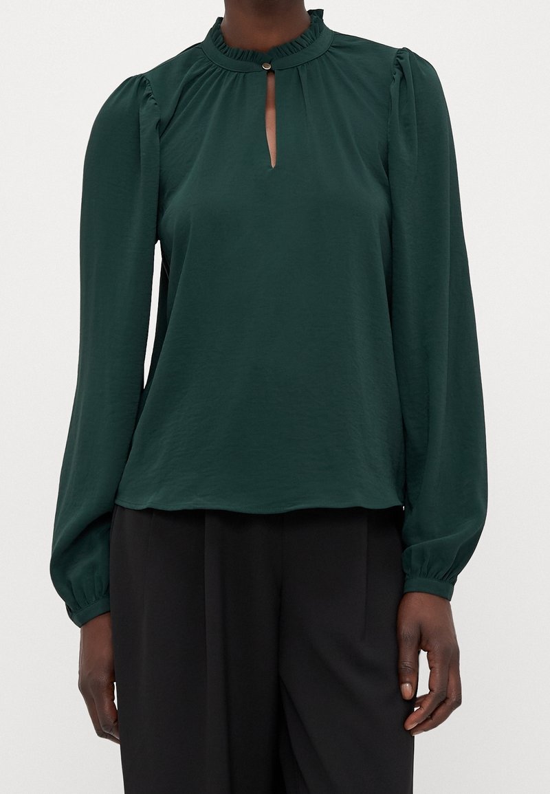 Blouse à manches longues vert foncé avec une petite ouverture en forme de goutte sur le devant et un col à volants, portée avec un pantalon noir ample.