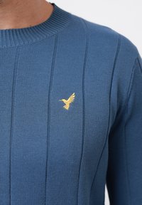 Niebieski sweter robiony na drutach z prążkowaną teksturą i pionowymi paskami; mała złota emblemat ptaka na lewej piersi.