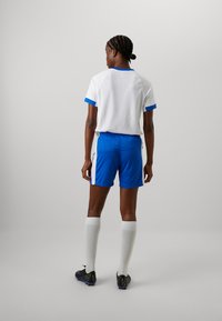 Jersey atlético blanco con acentos azules, combinado con pantalones cortos azules. Presenta textura acanalada, calcetines blancos y zapatos de fútbol negros.