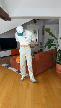 Άτομο που φοράει λευκό μπλουζάκι Adidas και ανοιχτόχρωμο παντελόνι, στέκεται εσωτερικά κοντά σε μια καφέ καναπέ και ένα μεγάλο φυτευτό φυτό.