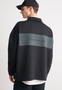 Felpa nera oversize con un pannello rettangolare grigio sul retro, con la scritta "Calvin Klein Jeans" in testo nero. Camicia bianca visibile sotto.