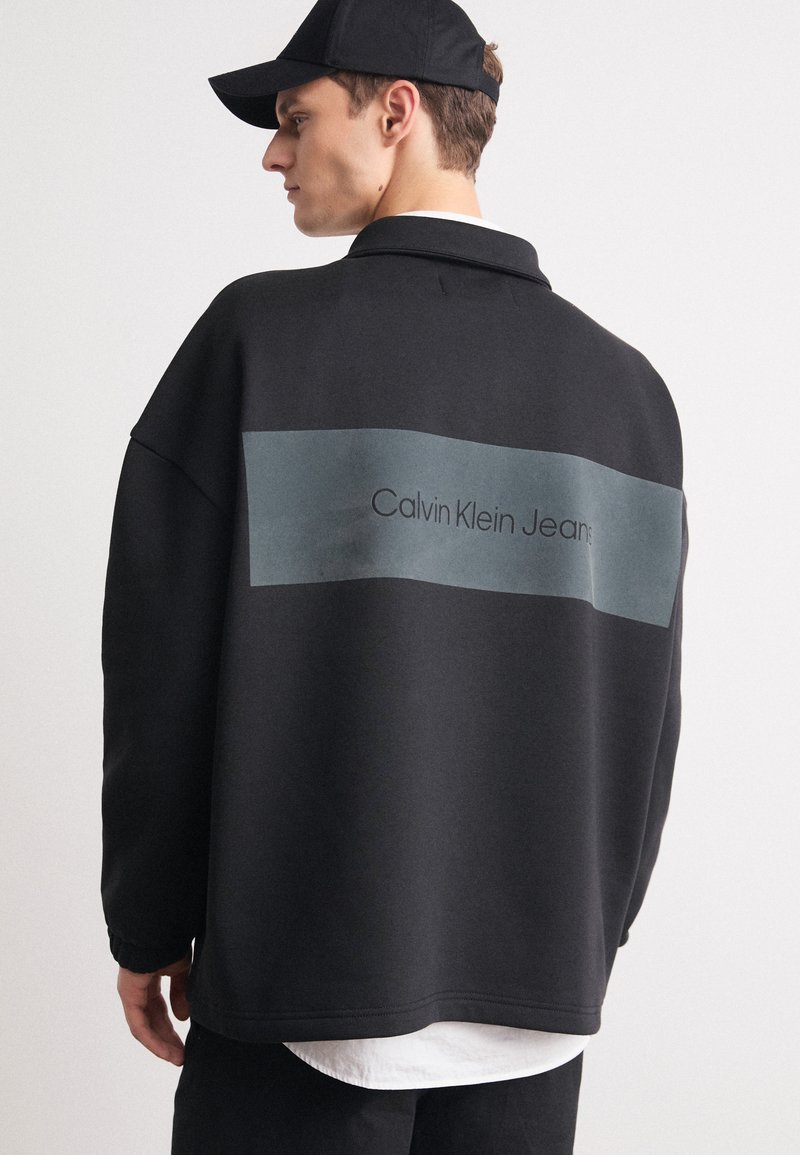 Felpa nera oversize con un pannello rettangolare grigio sul retro, con la scritta "Calvin Klein Jeans" in testo nero. Camicia bianca visibile sotto.