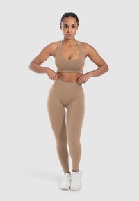 Hellbrauner Sport-BH und hoch taillierte Leggings aus glattem, elastischem Stoff. Weiße Sportschuhe komplettieren das Sportoutfit.
