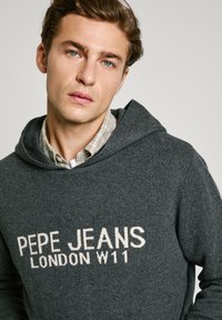 Sudadera gris con "PEPE JEANS LONDON W11" en texto blanco. Material suave con un corte relajado y capucha con cordón. Camisa de cuadros sutil por debajo.