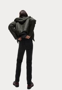 Olivově zelená bunda s kapucí a strukturovaným designem, nošená přes černý pletený svetr, kombinovaná s přiléhavými černými džíny a tmavými botami.