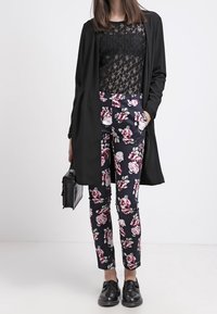 Černý dlouhý cardigan přes průhledný černý krajkový top, doplněný námořnickými květinovými kalhotami. Model nosí černé boty a nese malou tašku.