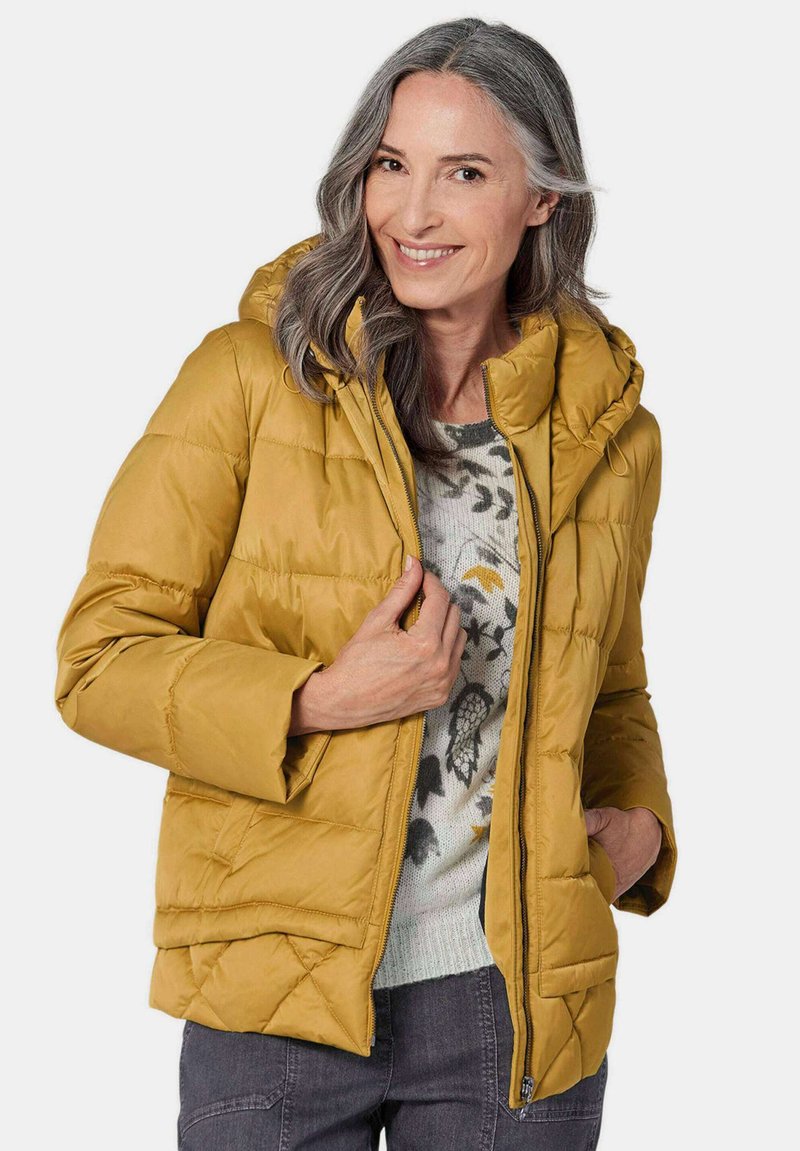Gelbe Steppjacke mit Kapuze, gestepptem Design, Reißverschluss, Seitentaschen und einem lockeren Schnitt, getragen über einem gemusterten Pullover.