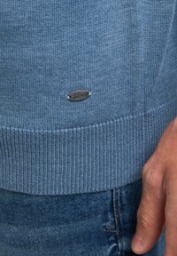 Blauwe gebreide trui met geribbelde manchetten en zoom, voorzien van een klein metalen logo-accent. Close-up van de trui en denim stof.