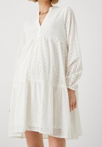 Weißes Spitzenkleid mit gestuftem Design, langen Ärmeln und V-Ausschnitt. Es besteht aus einem leichten Stoff mit komplexen Ausschnittmustern.