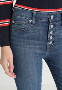 Högmidjade blå denimjeans med metallknappsstängning, fem-fickorsdesign och en lätt urblekt textur. Närbild av midjan och framsidan.