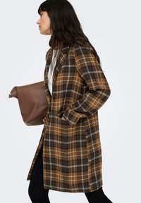 Palton maro în carouri, cu croială lejeră, reveri mari și două buzunare laterale. Asortat cu o geantă maro și pantaloni închiși la culoare.